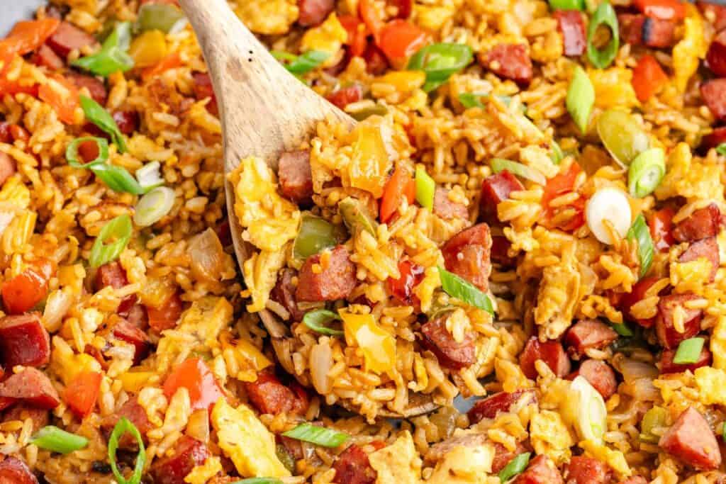 Close-up of fried rice with sausage, eggs, peppers, and green onions being stirred with a wooden spoon.