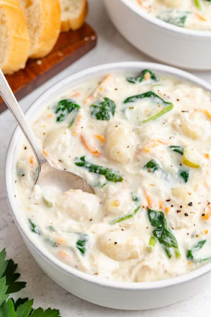 Olive Garden chicken gnocchi soup with spinach and carrots in a bowl, spoon inside, bread slices in the background.