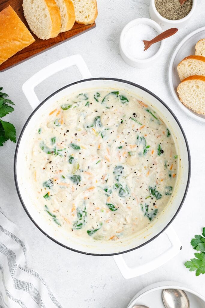 White pot of gnocchi chicken soup with spinach and carrots, surrounded by sliced bread, salt, pepper, and parsley.