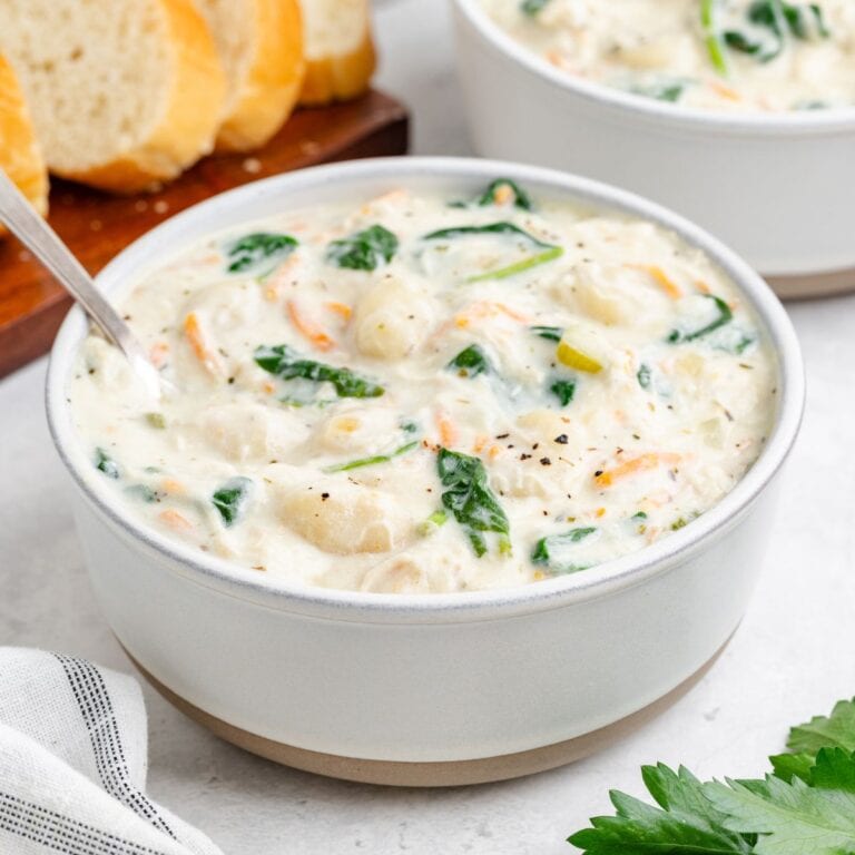 Creamy chicken gnocchi soup with spinach, and herbs in a bowl, spoon inside; sliced bread seen behind.