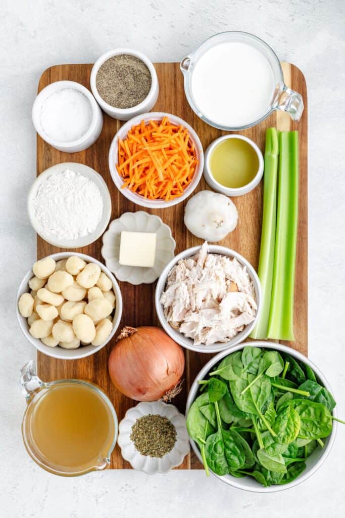 Wooden board with gnocchi, shredded chicken, spinach, vegetables, flour, butter, oil, milk, broth, and seasonings.