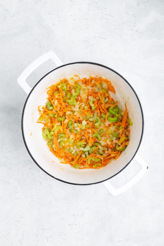 White pot with sautéed chopped onions, celery, and carrots on a light gray surface.