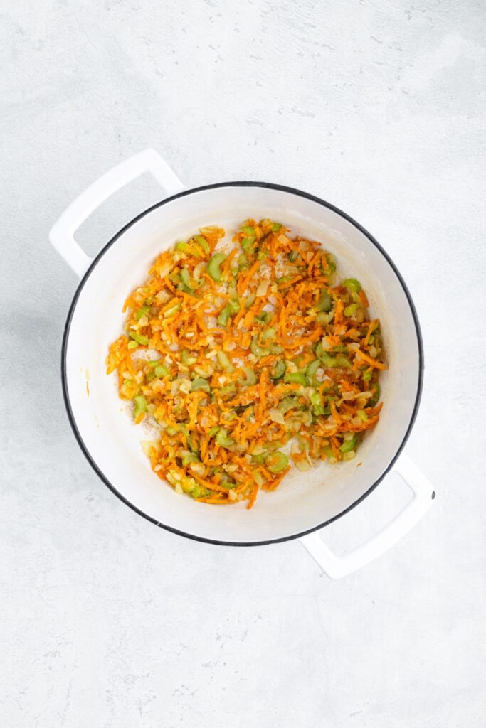 Chopped onions, celery, and carrots sauté in a white pot on a light surface.