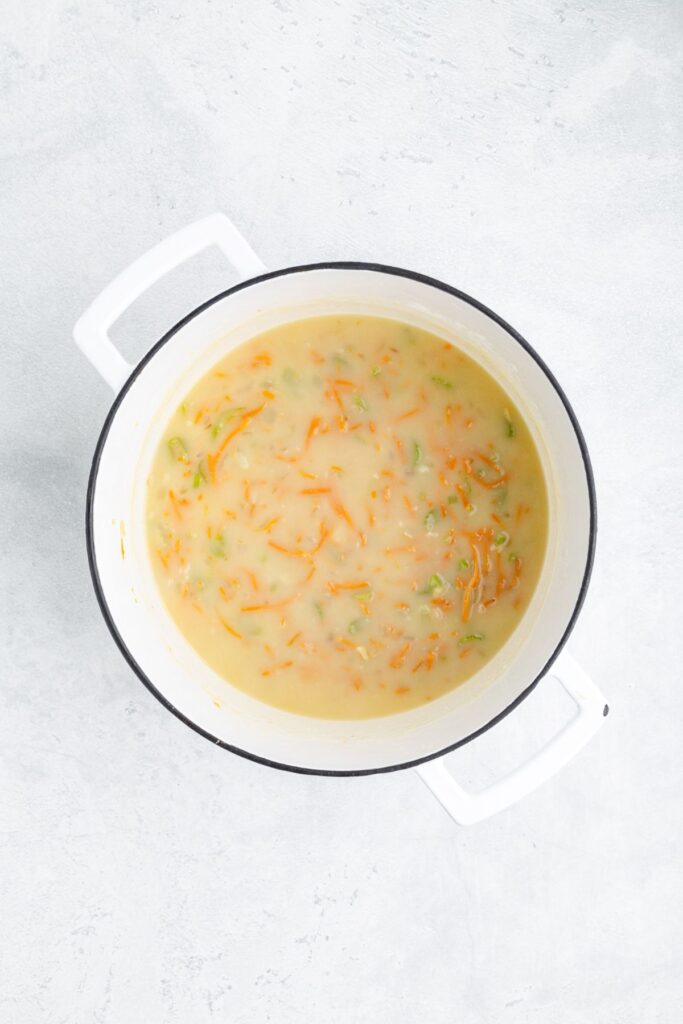 Creamy soup with carrots and green vegetables in a white pot, seen from above on a light surface.