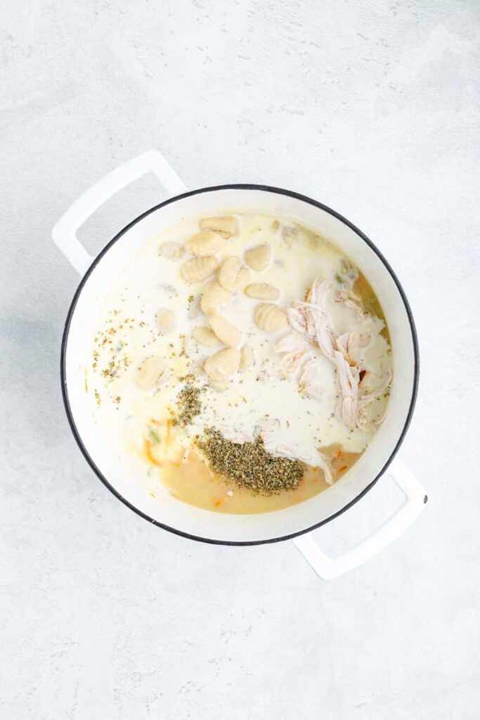 White pot filled with gnocchi, shredded chicken, herbs, broth, and cream sits on a light-colored surface.