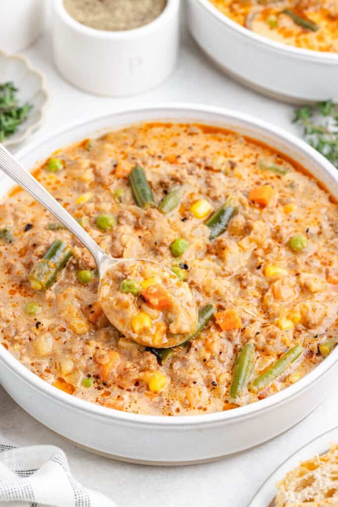 A bowl of creamy cottage pie soup with a spoon, showing corn, carrots, peas, and green beans.