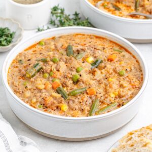A white bowl of creamy shepherd's pie soup with carrots, green beans, peas, corn, and herbs on a light surface.