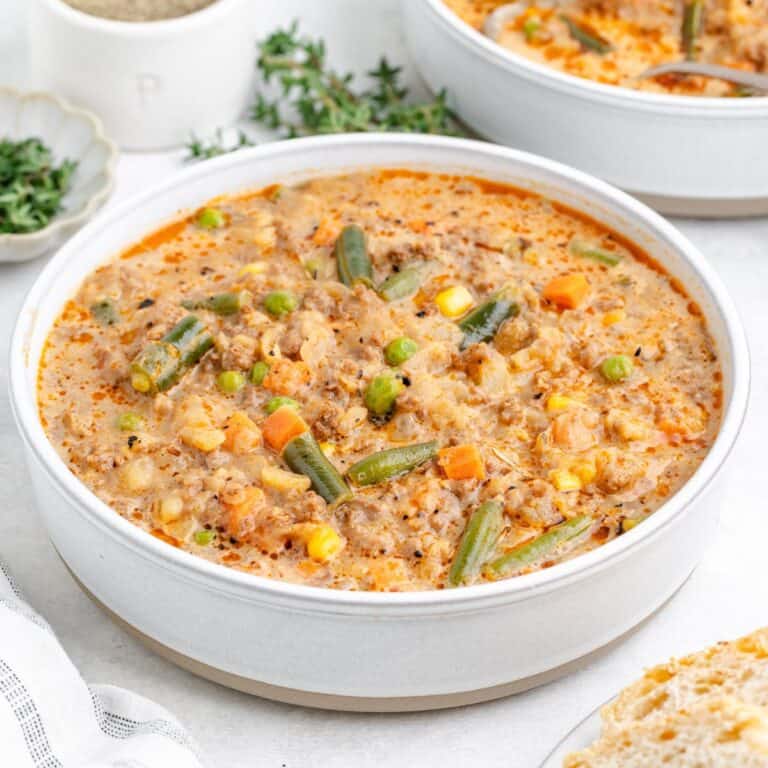 A white bowl of creamy shepherd's pie soup with carrots, green beans, peas, corn, and herbs on a light surface.