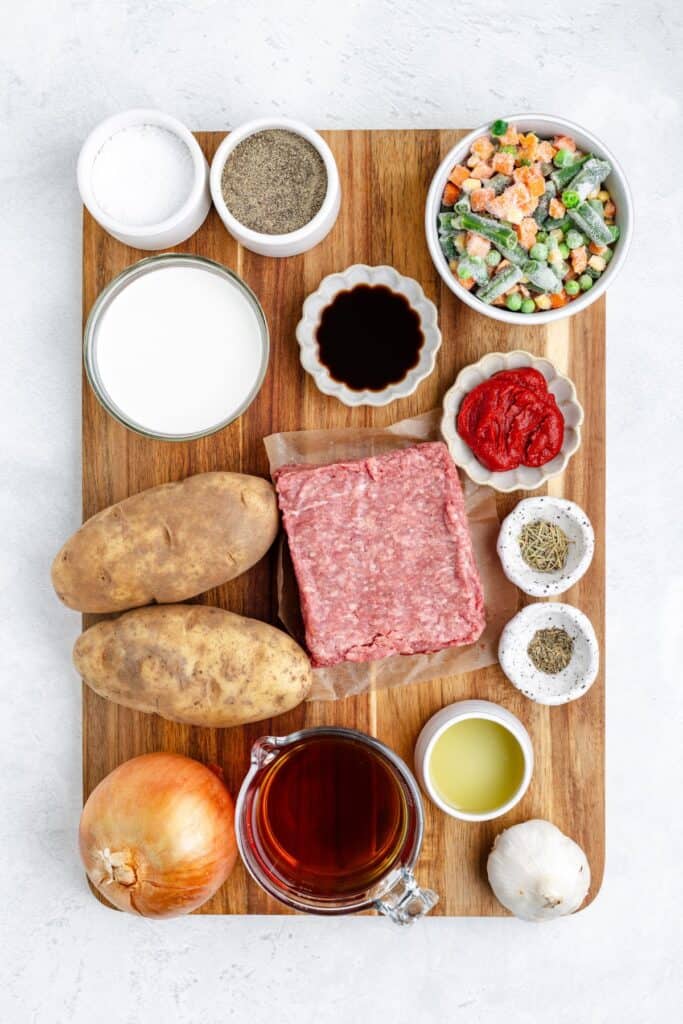 A wooden board topped with ground beef, potatoes, vegetables, milk, broth, tomato paste, oil, sauces, and spices.