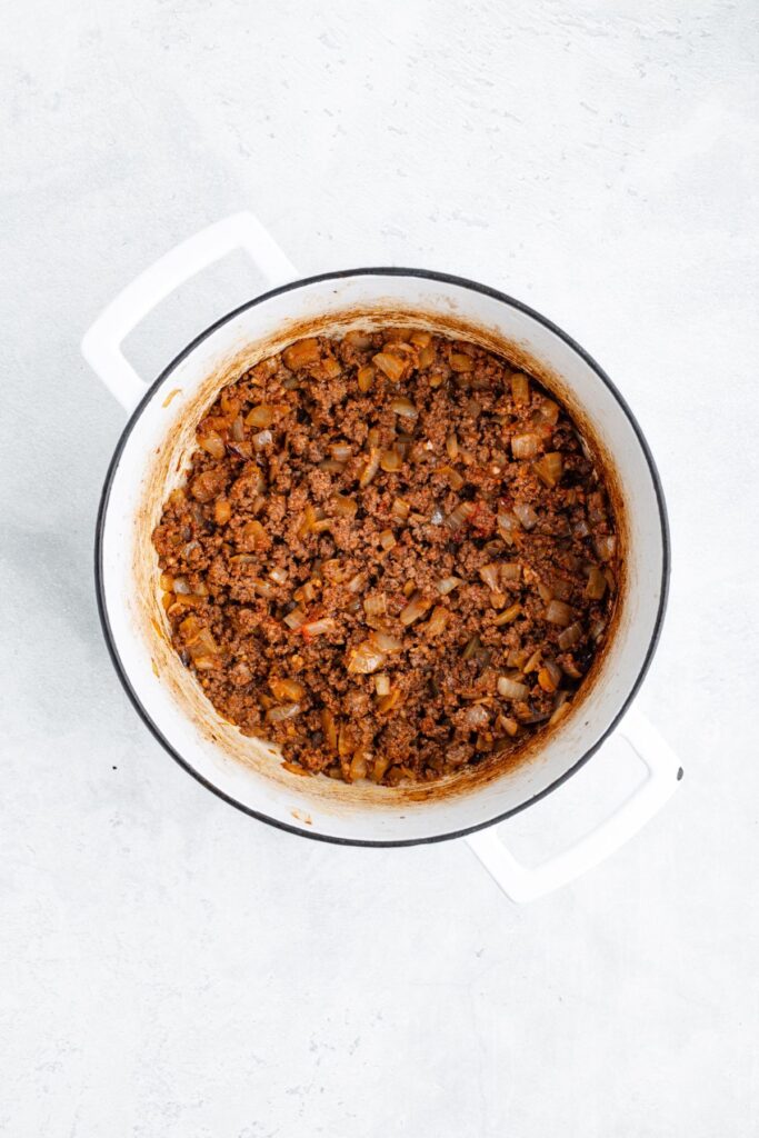 Cooked ground beef with diced onions and seasonings in a white pot, seen from above on a light surface.