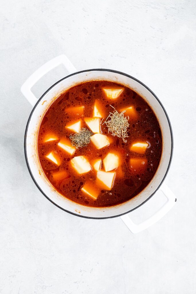 Red broth with potato chunks and dried herbs in a white pot, viewed from above on a light background.
