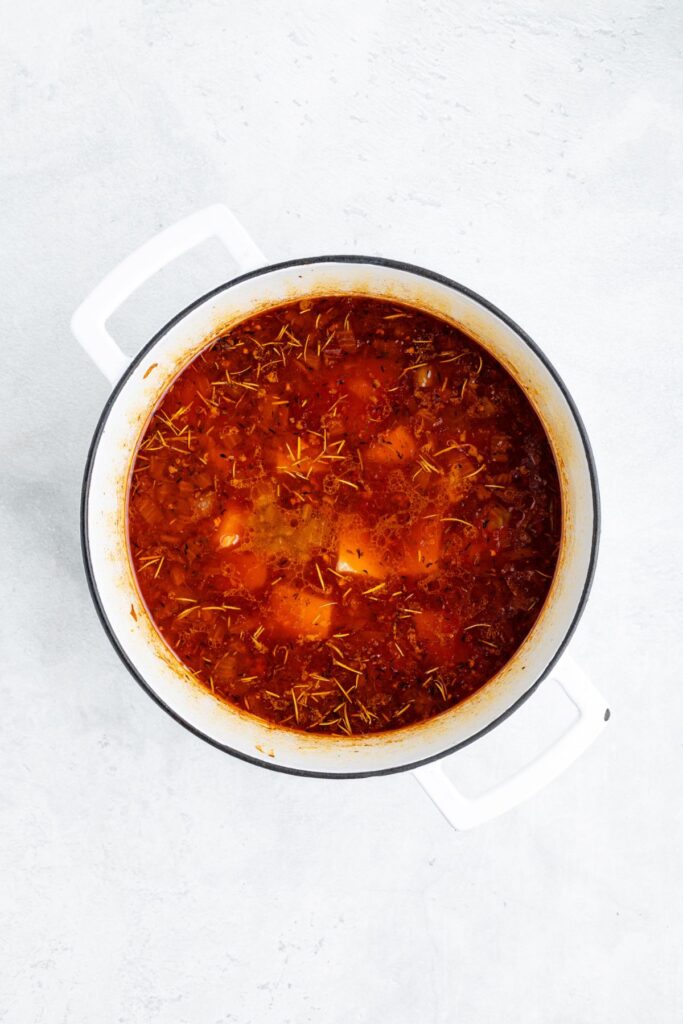 White pot with chunky red tomato stew topped with herbs, seen from above on a light-colored surface.