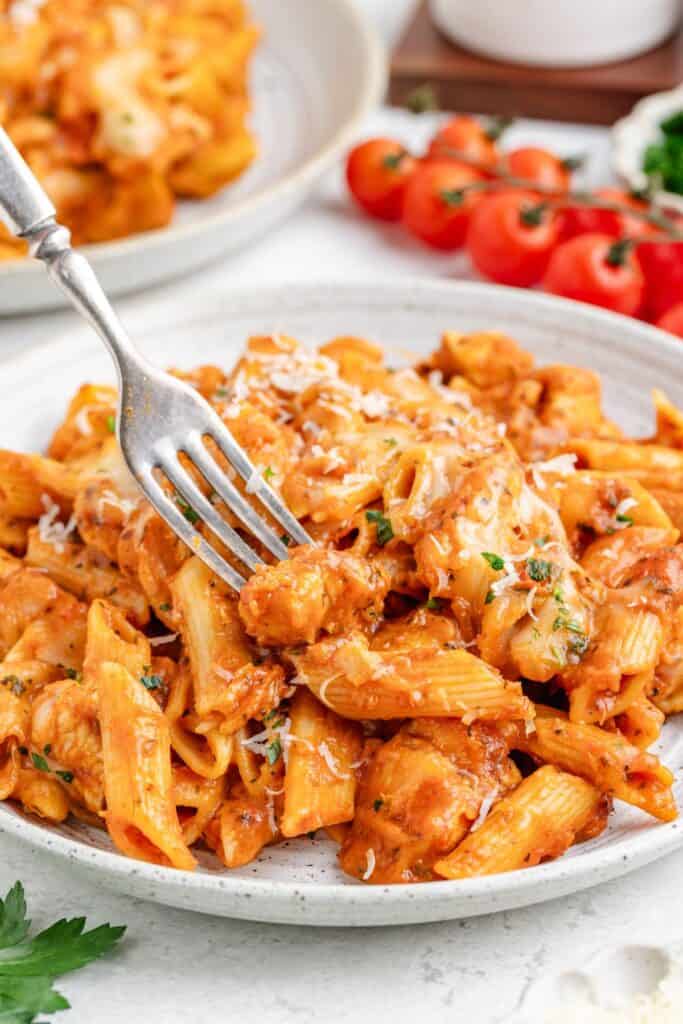 Chicken parmesan pasta with tomato sauce, chicken, cheese, and herbs on a plate, fork and tomatoes in background.