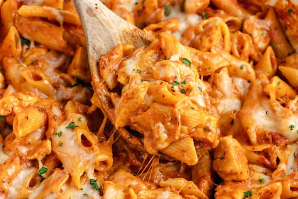 One pan chicken parmesan pasta with melted cheese and tomato sauce, served with a wooden spoon, close-up view.