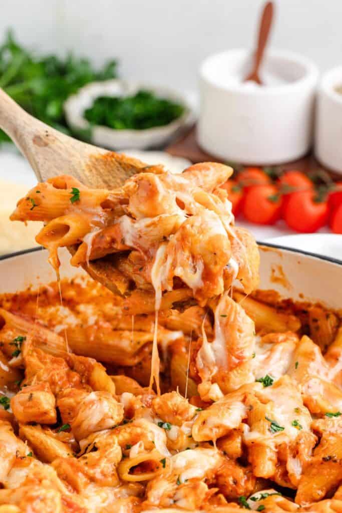 Wooden spoon scoops cheesy chicken parmesan pasta, with melted cheese stretch and herbs on top.