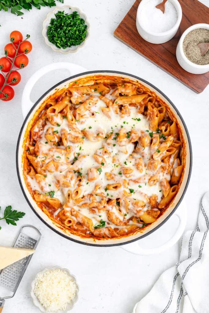 One pot chicken parmesan pasta in tomato sauce, melted cheese, and herbs on white surface with fresh ingredients.