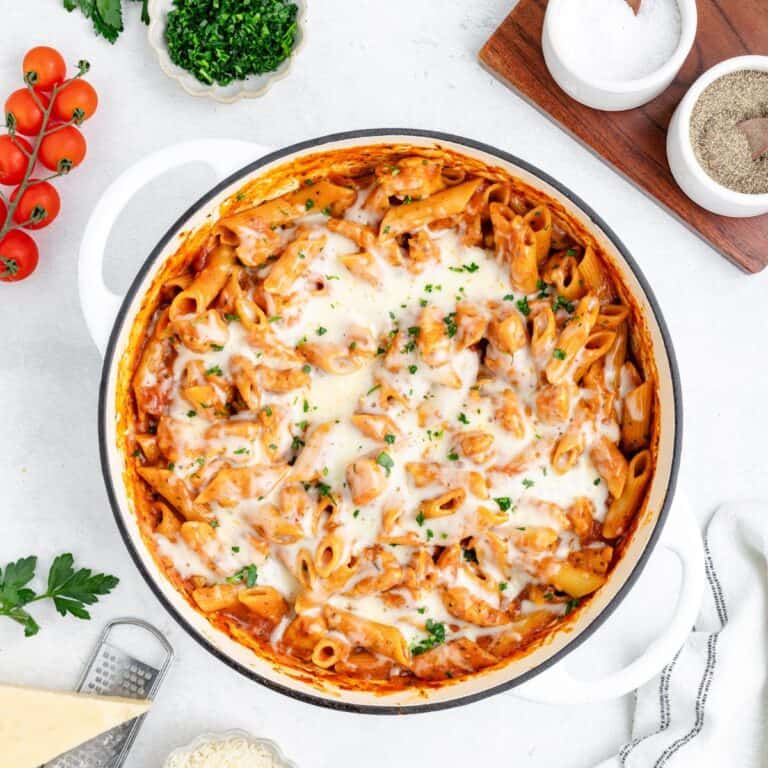 One pot chicken parmesan pasta with melted cheese in a white pot, surrounded by fresh herbs and cherry tomatoes.