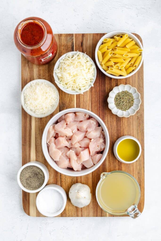 Wooden board with bowls of diced chicken, penne pasta, cheeses, spices, broth, and a jar of tomato sauce.
