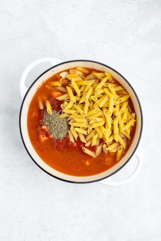 White pot containing uncooked penne, tomato sauce, chicken, seasonings, and broth on a light surface.