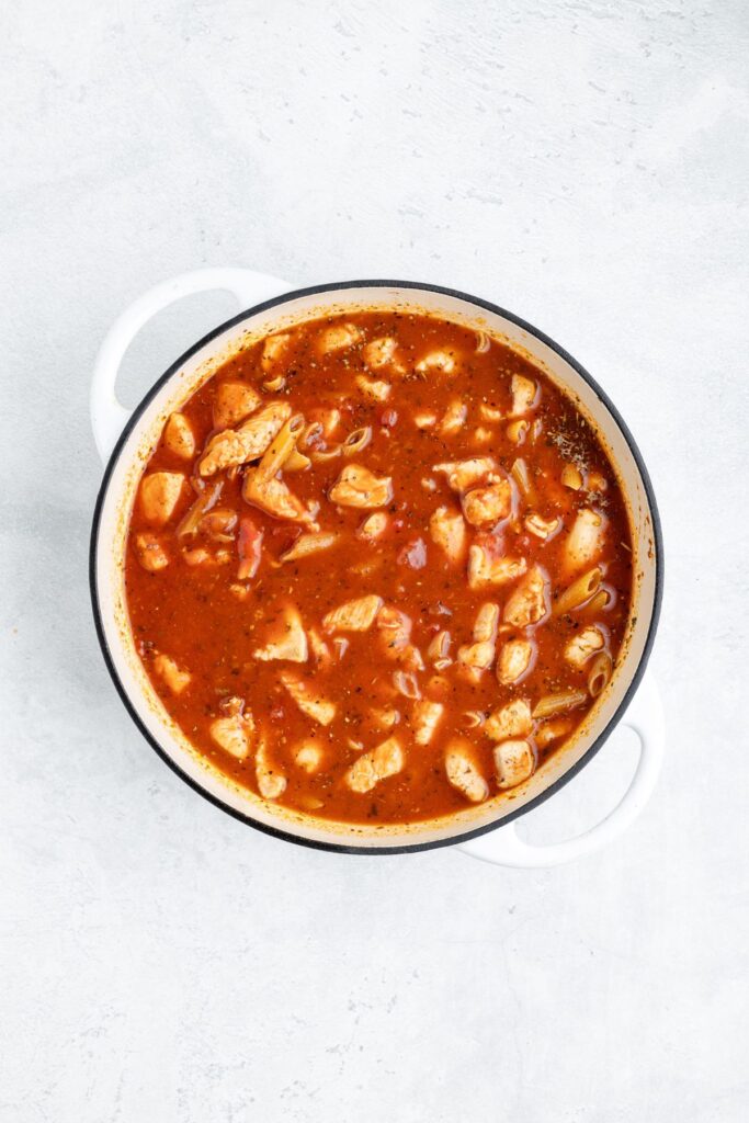 White pot with tomato soup, chicken chunks, and pasta sits on a light-colored surface.