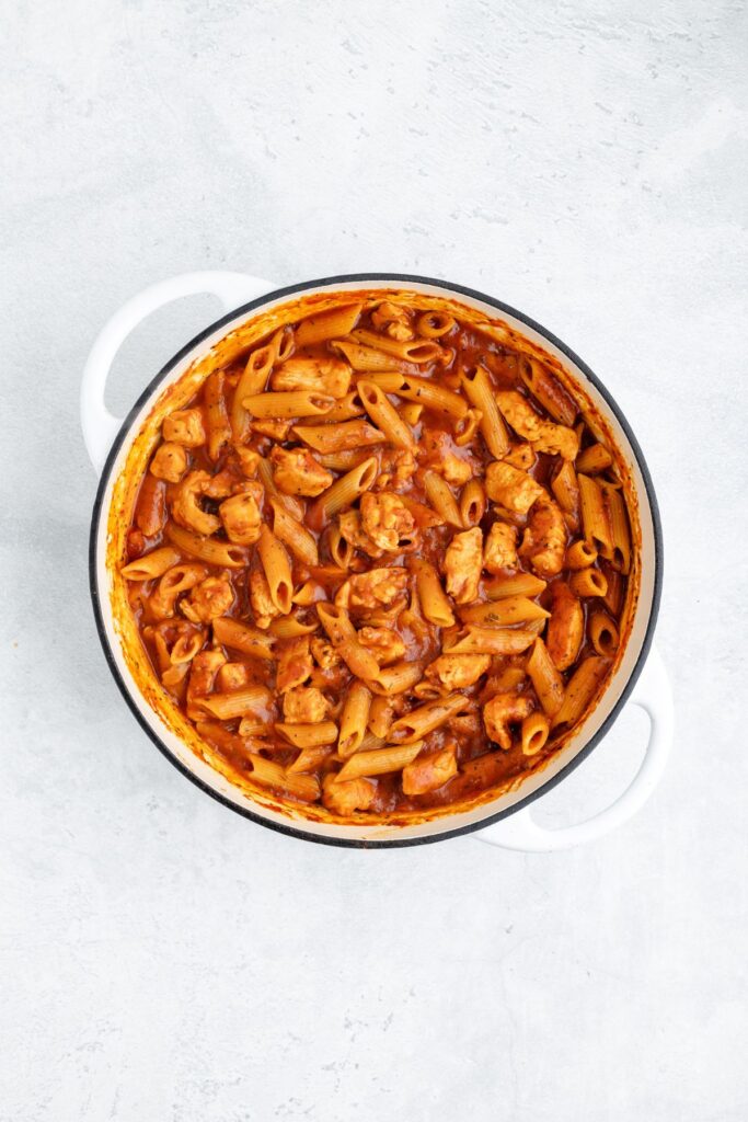 White pot with penne pasta, chicken pieces, and tomato sauce seen from above on a light surface.