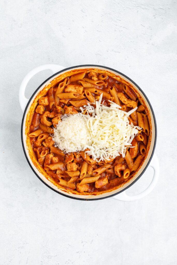 White pot with penne pasta, chicken in tomato sauce, and shredded cheese, seen from above on a light surface.