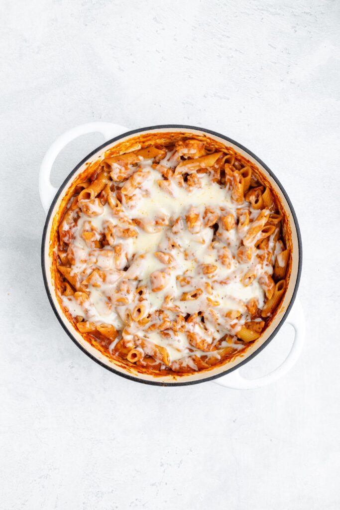Baked pasta with tomato sauce and melted cheese in a round white casserole dish on a pale surface.