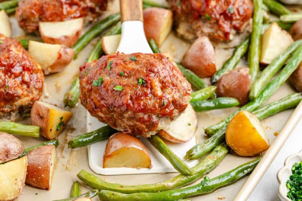 Sheet pan mini meatloaf, with green beans, roasted potatoes, and a spatula lifting one portion.