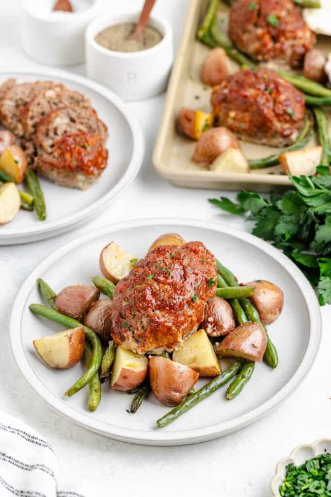 Plate of mini meatloaf, roasted green beans, and red potatoes with extra servings and seasonings in the background.
