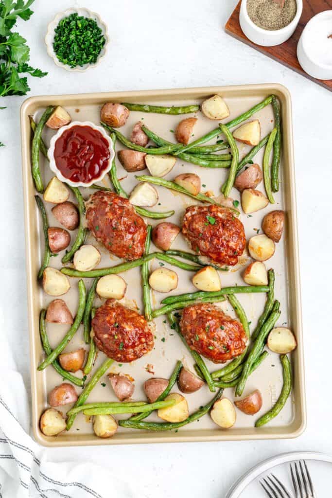 Sheet pan with four mini meatloaves, green beans, diced potatoes, and a small dish of ketchup.