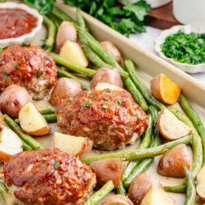 Sheet pan mini meatloaves with green beans, and roasted potatoes, topped with herbs; sauce bowls in background.