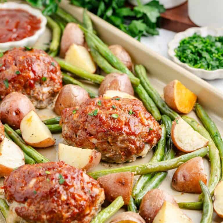 Sheet pan mini meatloaves with green beans, and roasted potatoes, topped with herbs; sauce bowls in background.