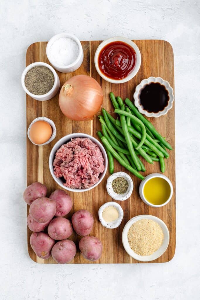 Ground meat, potatoes, green beans, onion, egg, condiments, breadcrumbs, and seasonings arranged on a wooden board.