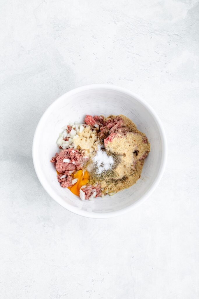 A white bowl with ground meat, chopped onion, garlic, breadcrumbs, seasonings, salt, and a raw egg on a light surface.