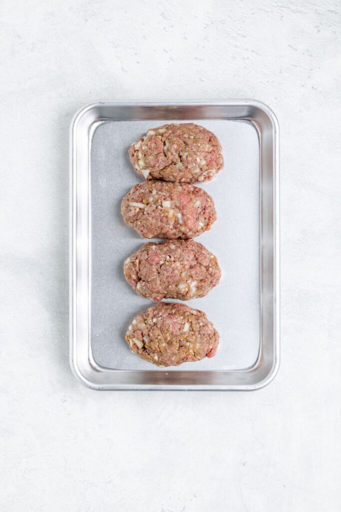 Metal tray with four raw oval ground meat patties mixed with onions on a light-colored surface.