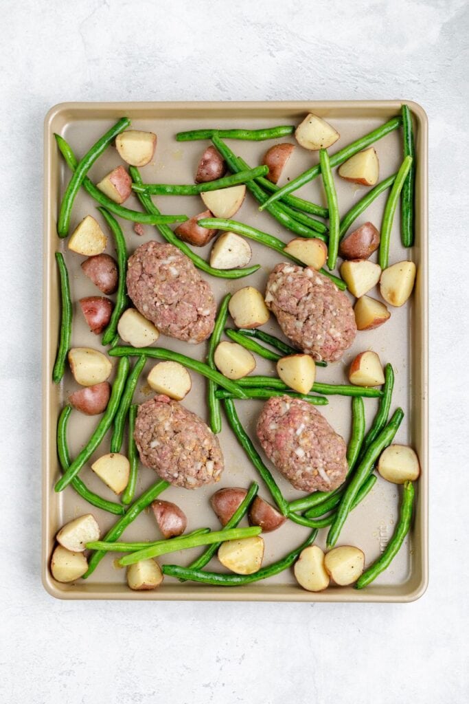 Baking sheet with four uncooked mini meatloaves, green beans, and red potato pieces arranged together.