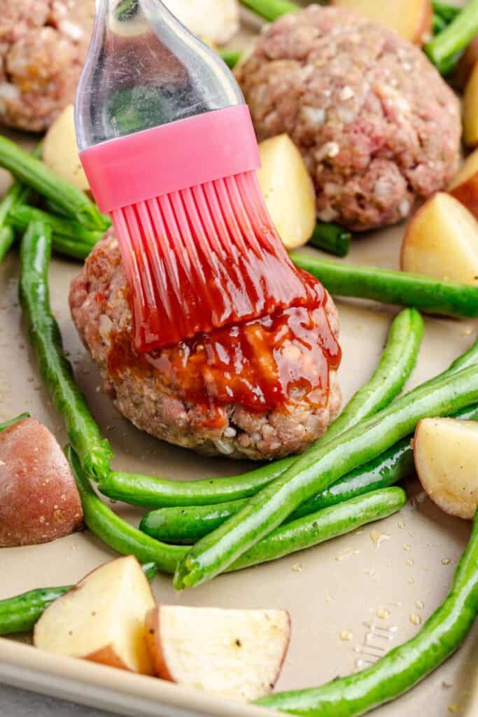 A silicone brush spreads sauce onto a raw meatloaf patty beside green beans and potatoes on a baking sheet.