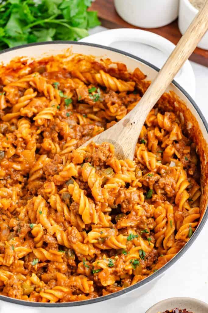 Sloppy Joe pasta in tomato meat sauce stirred with a wooden spoon, topped with fresh herbs in a pot.
