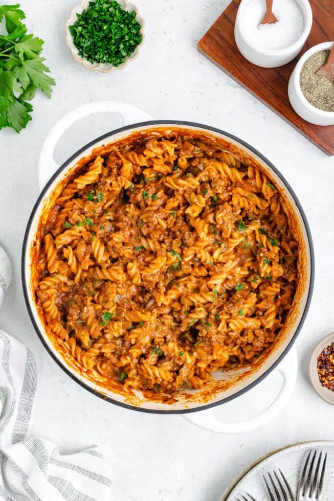 White pot of sloppy joe pasta in meat-tomato sauce with herbs, surrounded by condiments and a striped napkin.