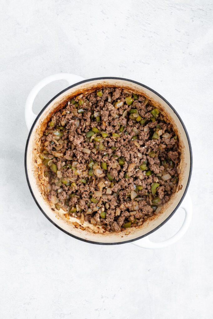 White pot with cooked ground beef, diced green bell peppers, and onions on a light gray surface.