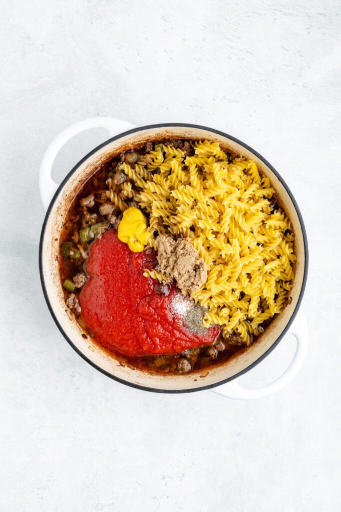 White pot holding separated cooked ground beef, uncooked rotini, tomato sauce, peas, mustard, brown sugar, and seasonings.