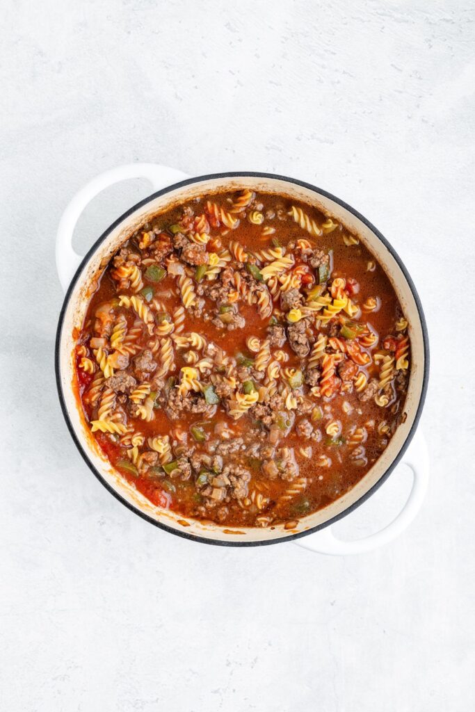 White pot with rotini pasta, ground beef, tomato sauce, and vegetables on a light gray surface.