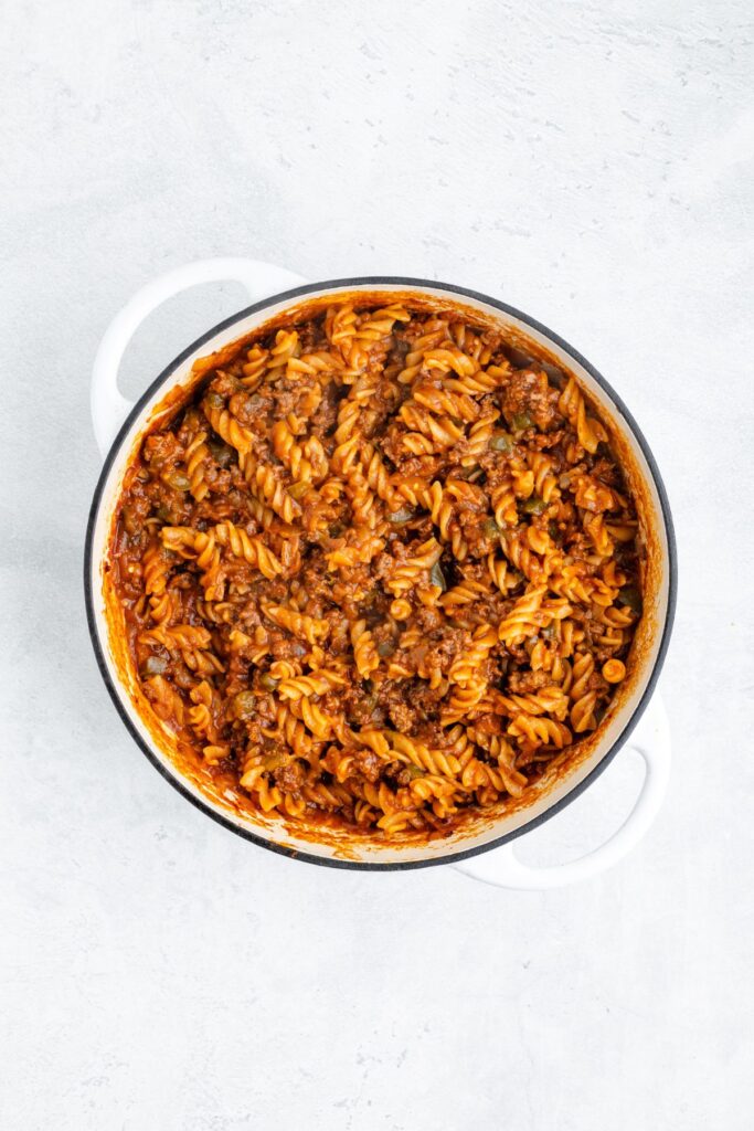 Cooked rotini pasta with ground beef and tomato sauce in a white pot, viewed from above on a light surface.