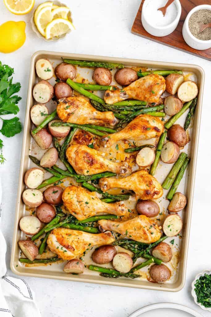 Oven baked chicken drumsticks, asparagus, and potatoes garnished with herbs; lemon slices and parsley nearby.