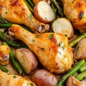 Roasted chicken drumsticks, red potatoes, and asparagus spears with herbs on a baking sheet.