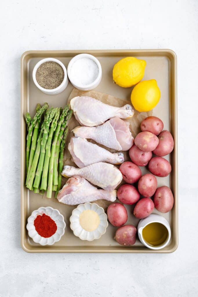 A baking sheet with grouped raw chicken drumsticks, asparagus, potatoes, lemons, oil, and seasonings.