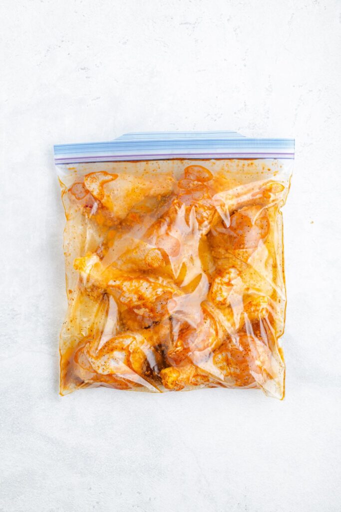 Raw chicken marinating in orange marinade inside a sealed zip-top bag on a light surface.