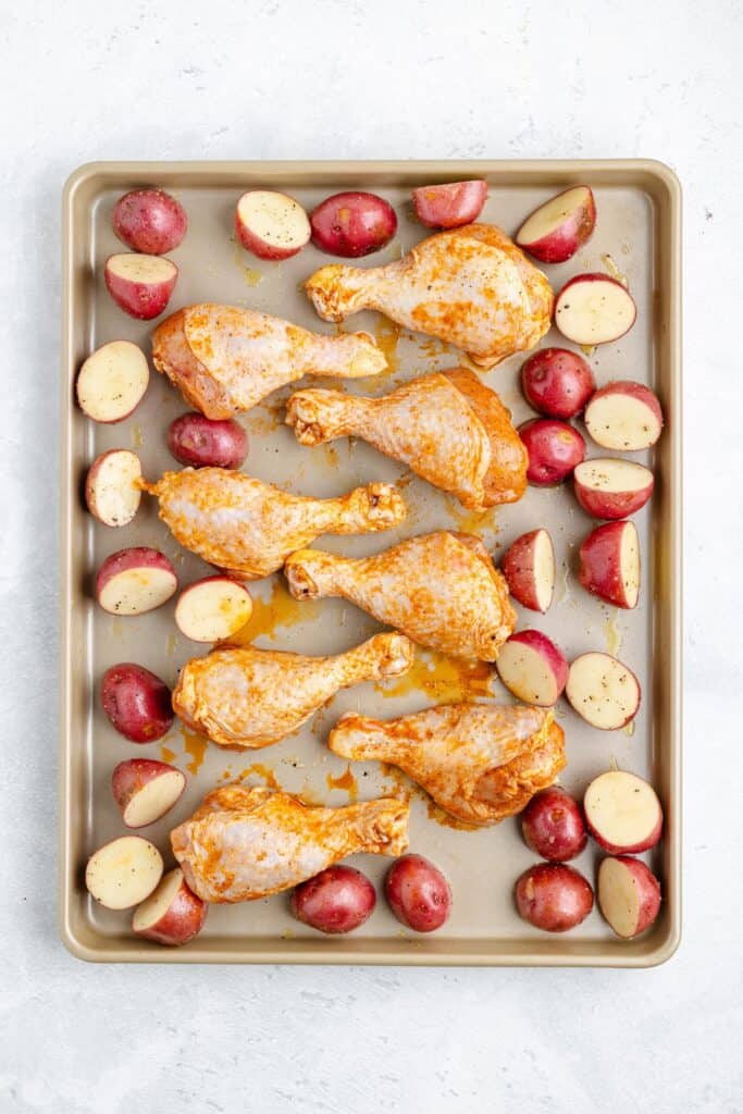 Seasoned raw chicken drumsticks and halved red potatoes placed on a baking sheet, prepared for roasting.