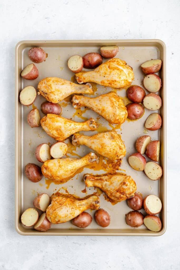 Six seasoned chicken drumsticks and halved red potatoes on a baking sheet, arranged for roasting.