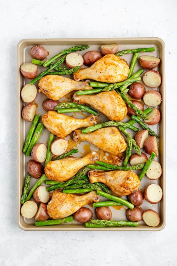 Seasoned chicken drumsticks, red potato halves, and asparagus spears on a light sheet pan.